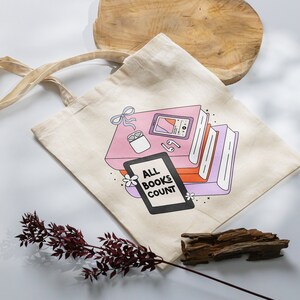 May include: A beige tote bag with a graphic of a stack of books, a phone, and earbuds. The graphic is in pink, orange, and purple. The text on the bag reads "All Books Count".
