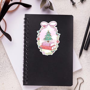 May include: A black spiral-bound notebook with a sticker featuring a Christmas tree, books, and the text "XMAS Book Club." The sticker is surrounded by a pink bow and floral elements. Other items include glasses, pens, and paper clips.