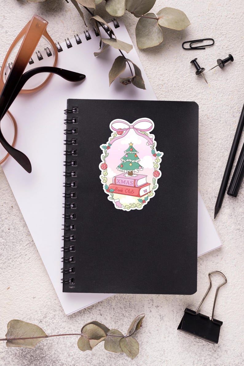 May include: A black spiral-bound notebook with a sticker featuring a Christmas tree, books, and the text "XMAS Book Club." The sticker is surrounded by a pink bow and floral elements. Other items include glasses, pens, and paper clips.
