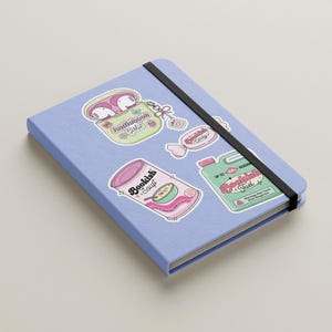 May include: A light blue notebook with a black elastic band, adorned with colorful stickers. The stickers feature illustrations of a can of soup, a container of earbuds, and candy, all with the theme of reading and books.
