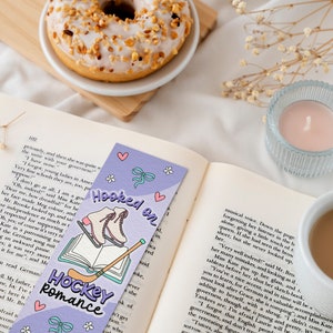 May include: A purple bookmark with a hockey stick and ice skates, with the text "Hooked on Hockey Romance". The bookmark is on top of an open book with a cup of coffee and a donut in the background.