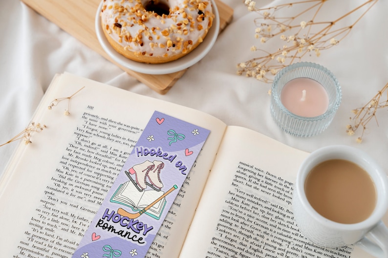 May include: A purple bookmark with a hockey stick and ice skates, with the text "Hooked on Hockey Romance". The bookmark is on top of an open book with a cup of coffee and a donut in the background.