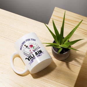 May include: White ceramic mug with a purple and white design featuring a book with a rose and a dagger, and the text "Falling for the Villain One Page at a Time".