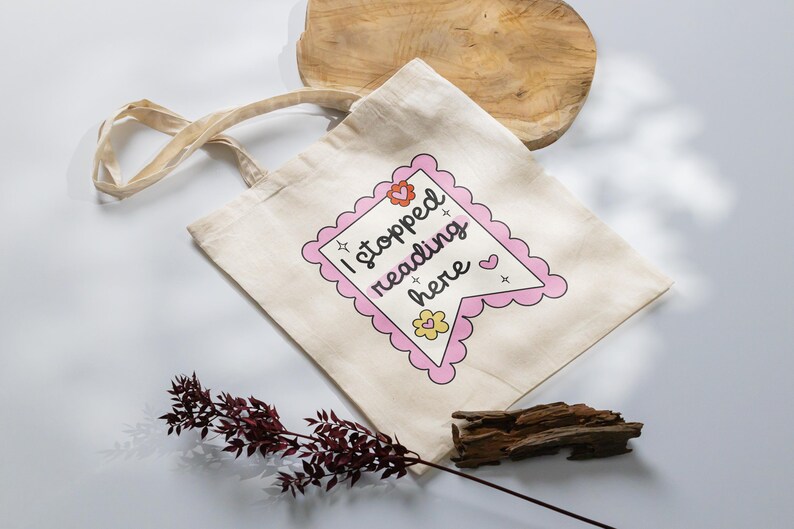 K&ouml;nnte beinhalten: Eine beige Tragetasche mit einem rosa und wei&szlig;en Banner, auf dem "Ich habe hier mit dem Lesen aufgeh&ouml;rt" steht, mit Herzen und Sternen.