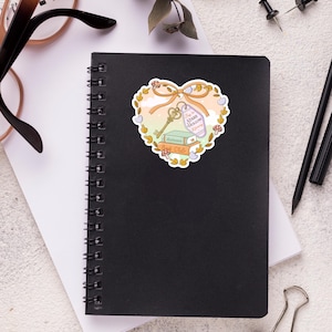 May include: A black spiral-bound notebook with a heart-shaped sticker. The sticker features a key, books, and the text "Stars Hollow Beginning". The sticker is surrounded by a floral design with a bow.