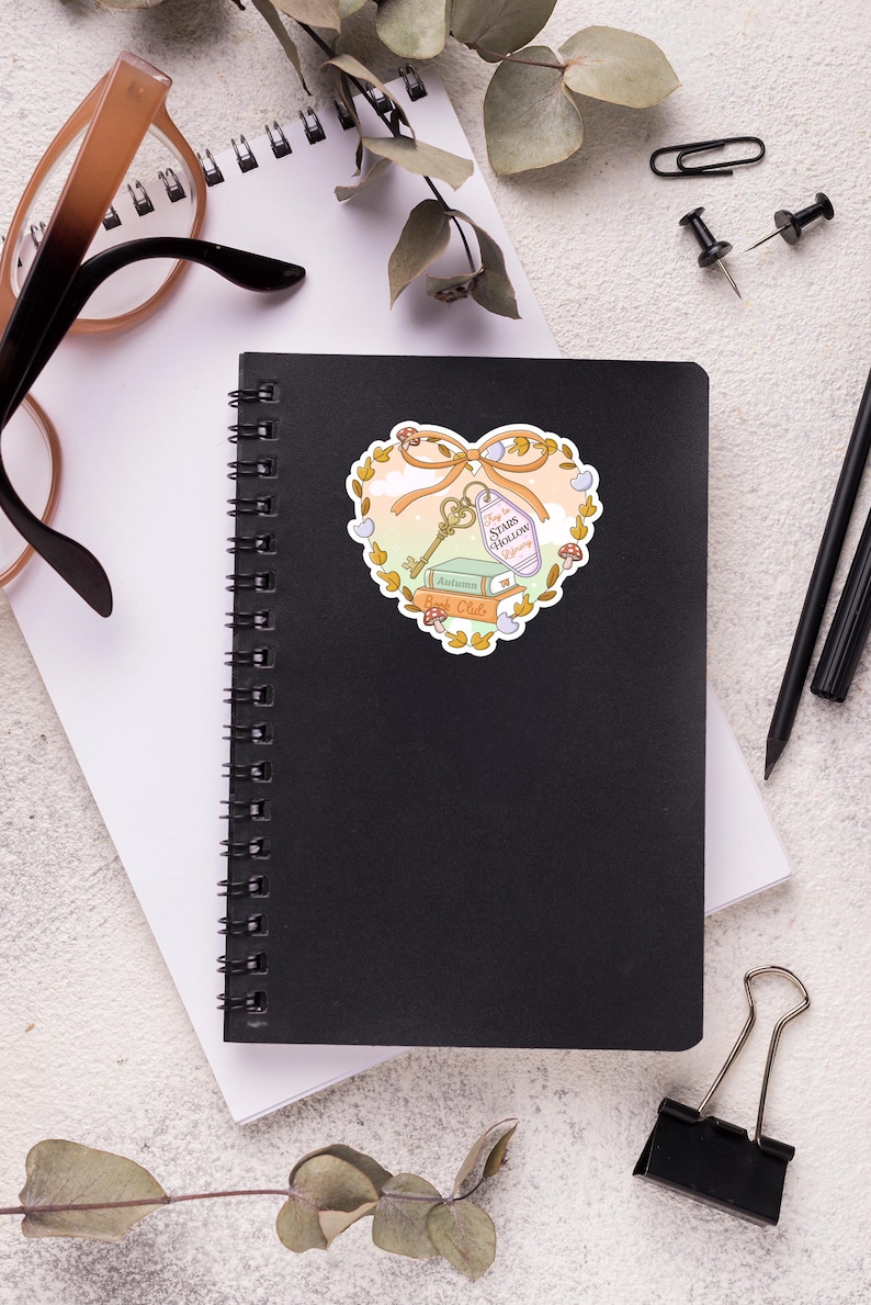May include: A black spiral-bound notebook with a heart-shaped sticker. The sticker features a key, books, and the text "Stars Hollow Beginning". The sticker is surrounded by a floral design with a bow.