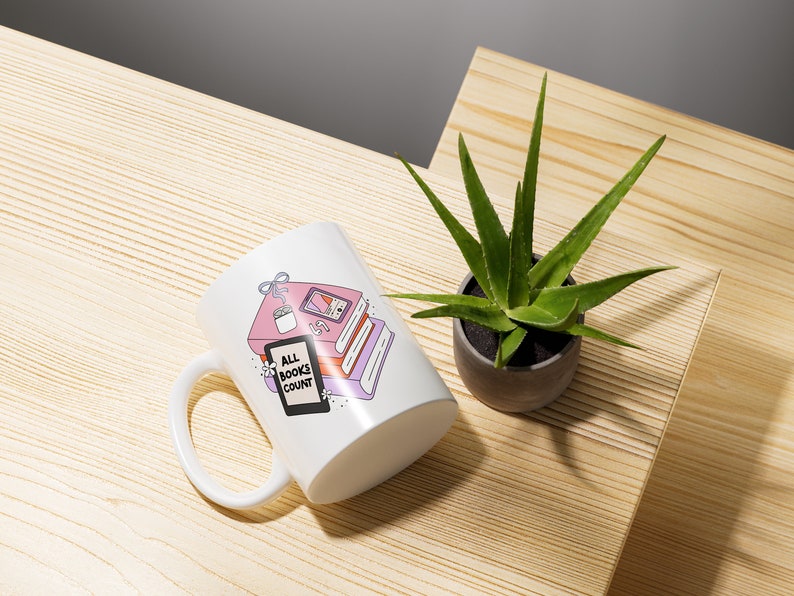 May include: White ceramic mug with a pink and purple illustration of a stack of books, a potted plant, and a phone. The text "All Books Count" is printed on the mug.
