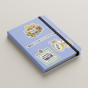 May include: A blue notebook with three stickers. One sticker is of a stack of books with a yellow ribbon and the text "Crime Scene Do Not Cross". Another sticker is of a candle with the text "Smells like she's reading a thriller". The third sticker is of a locket with the text "The Girl on the Train".