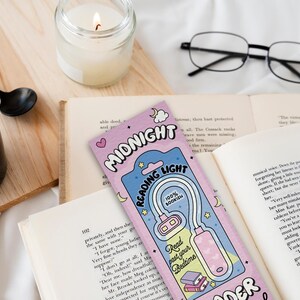 Puede incluir: Un marcap&aacute;ginas rosa y azul con una ilustraci&oacute;n de dibujos animados de una luz de lectura. El marcap&aacute;ginas dice "Midnight Reading Light" y "100% Bookish".