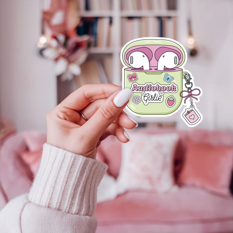 May include: A sticker featuring a cartoon illustration of a light green AirPod case with pink interior and white AirPods. The case has the text "Audiobook Girlie" and decorative elements like a bow, butterfly, flower, and strawberry.