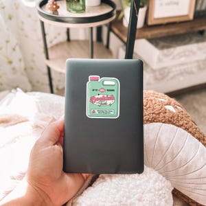 May include: A black laptop with a sticker on it that says "Bookish Fuel" in pink and green lettering. The sticker is shaped like a bottle with a pink lid and a white label. The sticker says "Up to Reading" on the label.