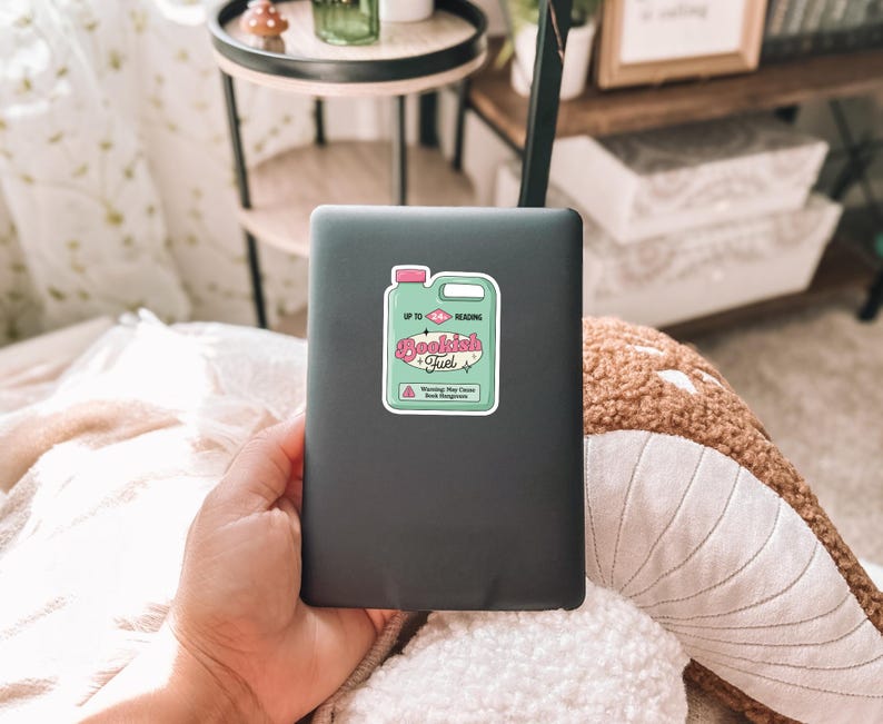 May include: A black laptop with a sticker on it that says "Bookish Fuel" in pink and green lettering. The sticker is shaped like a bottle with a pink lid and a white label. The sticker says "Up to Reading" on the label.
