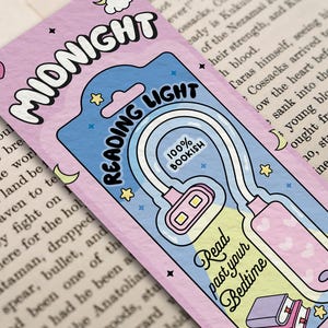 Puede incluir: Un marcap&aacute;ginas rosa y azul con una luz de lectura blanca adjunta. El marcap&aacute;ginas tiene una luna creciente, estrellas y el texto "MIDNIGHT READING LIGHT 100% BOOKISH". El texto "Read past your Bedtime" est&aacute; en la luz.