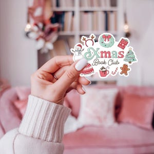 May include: A colorful sticker with the text "Xmas Book Club" and festive illustrations. The design includes a snowman, a wreath, a gift, a Christmas tree, a sweater, a mug of hot chocolate, a candy cane, and a gingerbread person.