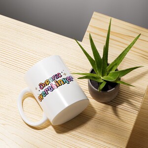 May include: White ceramic mug with a colorful graphic that says "Chronic overthinker". The mug is sitting on a wooden surface next to a small green plant in a gray pot.