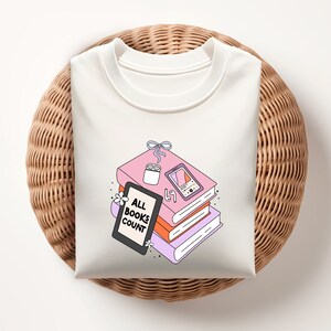 May include: White t-shirt with a graphic of a stack of books, a candle, a phone, and the text "ALL BOOKS COUNT".