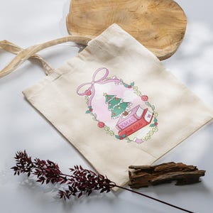 May include: A beige tote bag with a Christmas-themed design. The design features a Christmas tree, books, and the text "XMAS Book Club" inside a pink oval frame with a bow. The bag has beige handles.