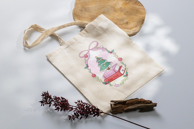 May include: A beige tote bag with a Christmas-themed design. The design features a Christmas tree, books, and the text "XMAS Book Club" inside a pink oval frame with a bow. The bag has beige handles.