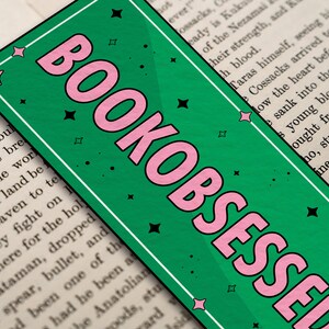 May include: A green bookmark with pink lettering that reads "BOOKOBSESSED". The bookmark has a white border and black stars.