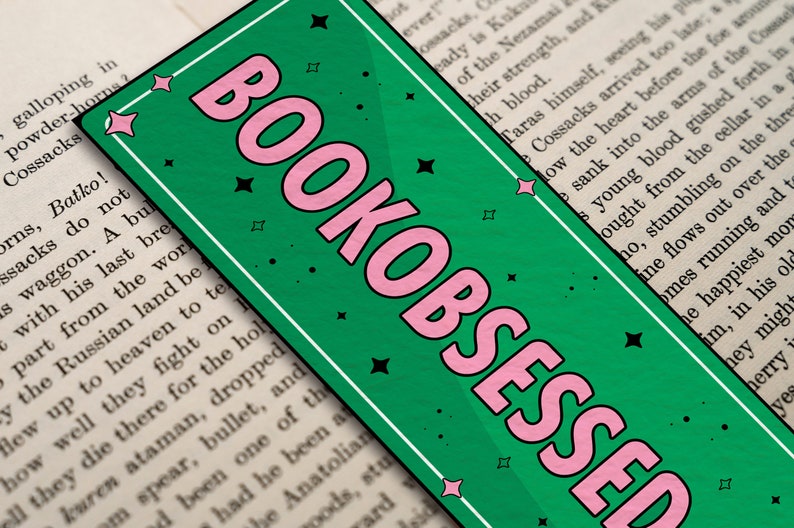 May include: A green bookmark with pink lettering that reads "BOOKOBSESSED". The bookmark has a white border and black stars.