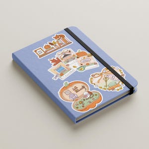 May include: A light blue notebook with a black elastic band, adorned with colorful stickers. The stickers feature autumn-themed illustrations, including pumpkins, leaves, and a fox, creating a cozy, seasonal aesthetic.