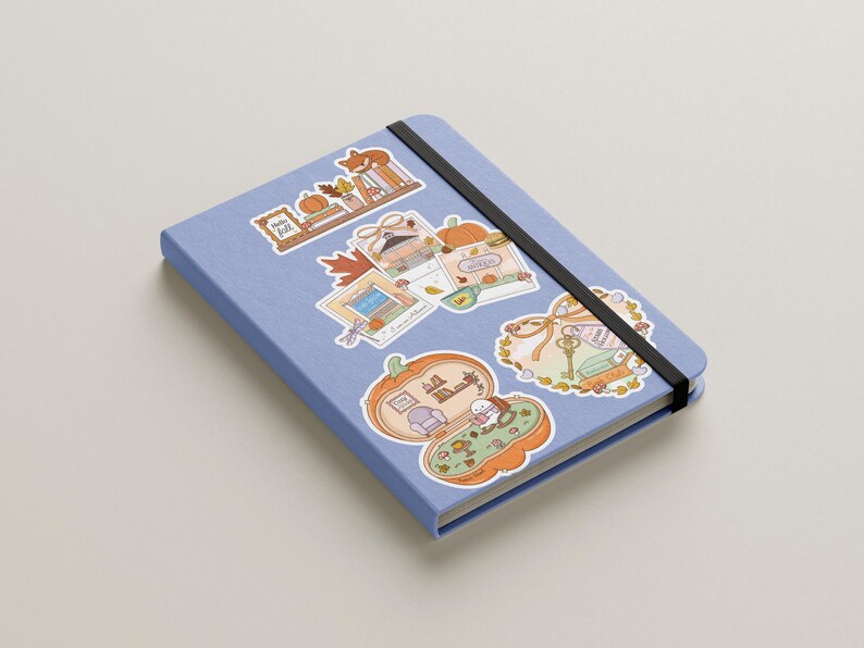 May include: A light blue notebook with a black elastic band, adorned with colorful stickers. The stickers feature autumn-themed illustrations, including pumpkins, leaves, and a fox, creating a cozy, seasonal aesthetic.
