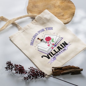 May include: A beige tote bag with a graphic of a book with a red rose and a dagger. The text on the bag reads "Falling for the Villain One Page at a Time."