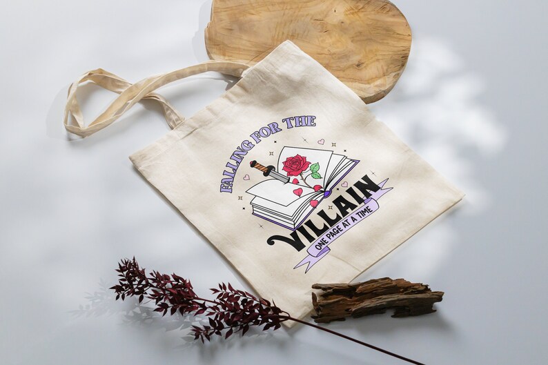May include: A beige tote bag with a graphic of a book with a red rose and a dagger. The text on the bag reads "Falling for the Villain One Page at a Time."