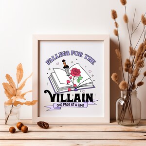 May include: A white framed print with a purple and pink illustration of an open book with a rose and a dagger sticking out of it. The text reads "Falling for the Villain, One Page at a Time."