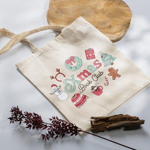 May include: A cream-colored tote bag with the text "Xmas Book Club" and festive illustrations. The design includes a snowman, gingerbread man, Christmas tree, and other holiday-themed graphics. The bag has two handles.