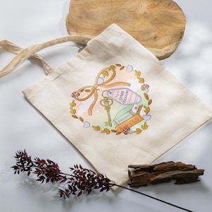 May include: Cream-colored tote bag with a whimsical design featuring a key, a bow, a hotel keychain that says "Stars Hollow", books, and mushrooms. The design is surrounded by a wreath of leaves and flowers. The bag has long handles.