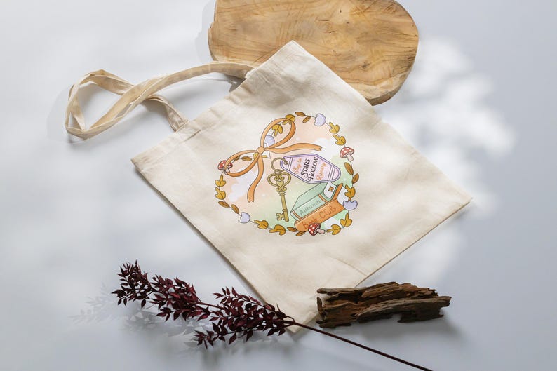 May include: Cream-colored tote bag with a whimsical design featuring a key, a bow, a hotel keychain that says "Stars Hollow", books, and mushrooms. The design is surrounded by a wreath of leaves and flowers. The bag has long handles.