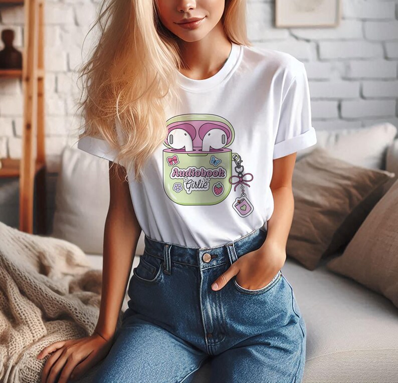 May include: White t-shirt featuring a cartoon graphic of a light green AirPods case with pink earbuds. The case has the text "Audiobook Girlie" and a small charm. The shirt is paired with blue jeans.