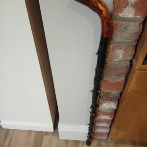 May include: A curved walking stick with a varnished brown handle and a black shaft. The shaft has small, dark protrusions. The stick leans against a white wall and a brick structure.