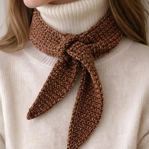 Puede incluir: Bufanda marrón tejida anudada al cuello, con extremos largos y puntiagudos. La bufanda se lleva sobre un jersey de cuello alto blanco. La textura tejida es visible, y el color es un tono cálido y terroso.