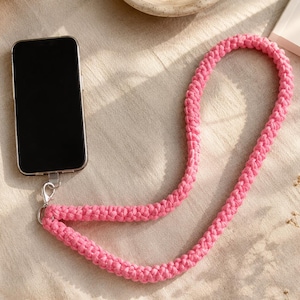 Può includere: Un cordino per telefono in corda intrecciata rosa brillante con chiusura a moschettone in metallo argentato. Il cordino è realizzato in un materiale tessile intrecciato spesso ed è collegato a uno smartphone nero tramite una piccola linguetta in plastica trasparente.