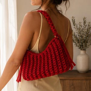 May include: A vibrant red crochet shoulder bag featuring a thick, braided strap and long, dangling fringe accents on the sides. The bag is constructed with a chunky, textured knit pattern and is secured with gold-toned metal hardware rings.