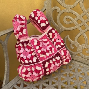 Può includere: Borsa tote all'uncinetto fatta a mano con un motivo a quadretti granny. La borsa presenta un mix di quadrati rosa, rossi e bianchi, con manici abbinati. La borsa è appoggiata su una panchina di metallo.