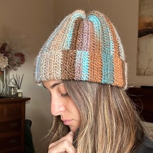 Può includere: Un berretto lavorato a mano a coste, in una miscela di colori tra cui beige, marrone, blu e lavanda. Il cappello è ripiegato sul bordo e sembra essere fatto di un filato morbido e strutturato. Un accessorio elegante per il freddo.