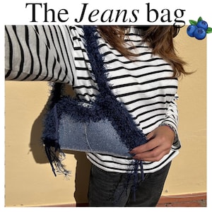 Può includere: Borsa a tracolla in denim con finiture e cinturino blu scuro con frange. La borsa è rettangolare e presenta la scritta "The Jeans bag" in grassetto, in nero.