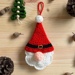 Può includere: Un ornamento di Babbo Natale all'uncinetto con un cappello rosso, una barba bianca e una cintura nera. L'ornamento è appeso a una superficie di legno con altre decorazioni natalizie.