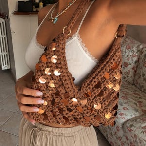 Può includere: Una borsa a spalla all'uncinetto marrone con tracolla intrecciata e paillettes circolari metalliche color rame applicate sull'esterno. La borsa presenta un'apertura a V ed è realizzata in filato spesso e testurizzato marrone scuro.