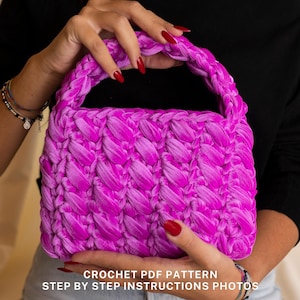 May include: A bright pink crocheted handbag with a braided handle. The bag is held by a person's hands. The text "CROCHET PDF PATTERN STEP BY STEP INSTRUCTIONS PHOTOS" is at the bottom of the image.