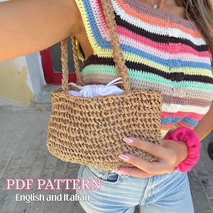 Può includere: Una borsa di paglia intrecciata di forma rettangolare con manici intrecciati. La borsa è tenuta da una persona che indossa un top all'uncinetto a righe colorate. Un elastico rosa è sul polso. Il testo "PDF PATTERN English and Italian" è in basso.