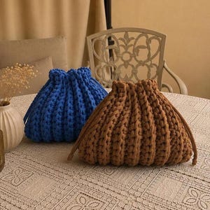 Può includere: Due pochette con coulisse realizzate in filato di lana spesso e voluminoso. Una pochette è di un blu reale vivace, mentre l'altra è di un caldo marrone terroso. Entrambe le borse presentano una chiusura superiore arricciata con cordini abbinati, creando un aspetto testurizzato e a coste sull'esterno di ogni pochette.