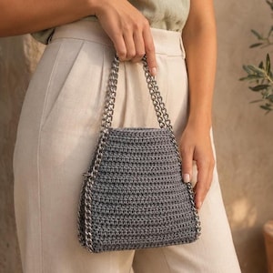 Può includere: Una borsa all'uncinetto grigio scuro di forma trapezoidale con un manico a catena in metallo color argento. La borsa presenta una struttura in filato intrecciato e viene tenuta per le cinghie a catena, mostrando un design strutturato e metallico.