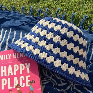 Può includere: Un cappello a secchiello all'uncinetto blu e crema con un motivo geometrico è appoggiato accanto a un libro intitolato "Happy Place" di Emily Henry. Il libro ha una copertina rosa con illustrazioni di persone. Il cappello è su un tessuto blu fantasia.
