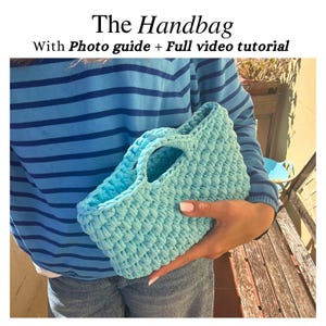 Può includere: Una borsa all'uncinetto azzurra con due manici. La borsa è tenuta da una persona che indossa una camicia a righe blu e jeans. Il testo "The Handbag With Photo guide + Full video tutorial" è nella parte superiore dell'immagine.