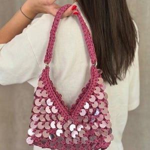 May include: A pink crocheted handbag with a sequined front. The bag has a V-shaped design and a braided handle. The sequins are circular and reflect light. The bag is held over a person's shoulder.