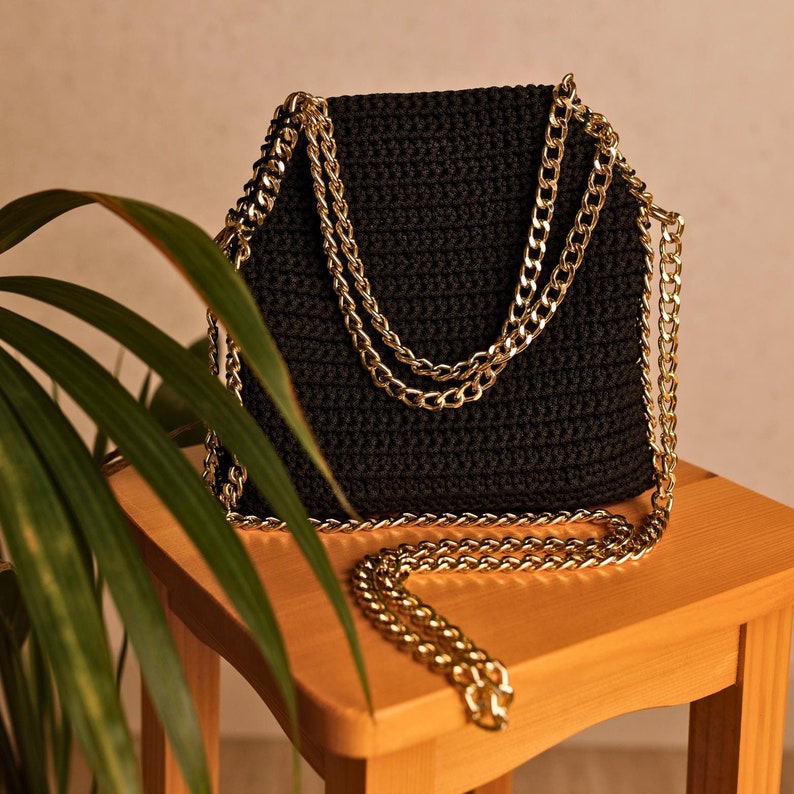 May include: A black crochet handbag with a gold chain strap. The bag is rectangular with a flap closure and a gold chain strap. The bag is sitting on a small wooden stool. A green plant is in the foreground.